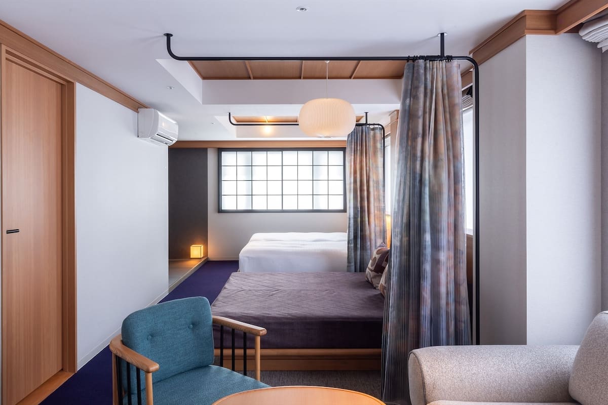 A compact hotel room with a bed, a chair, a small table, a window with curtains, and an air conditioning unit.