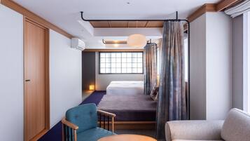 A compact hotel room with a bed, a chair, a small table, a window with curtains, and an air conditioning unit.