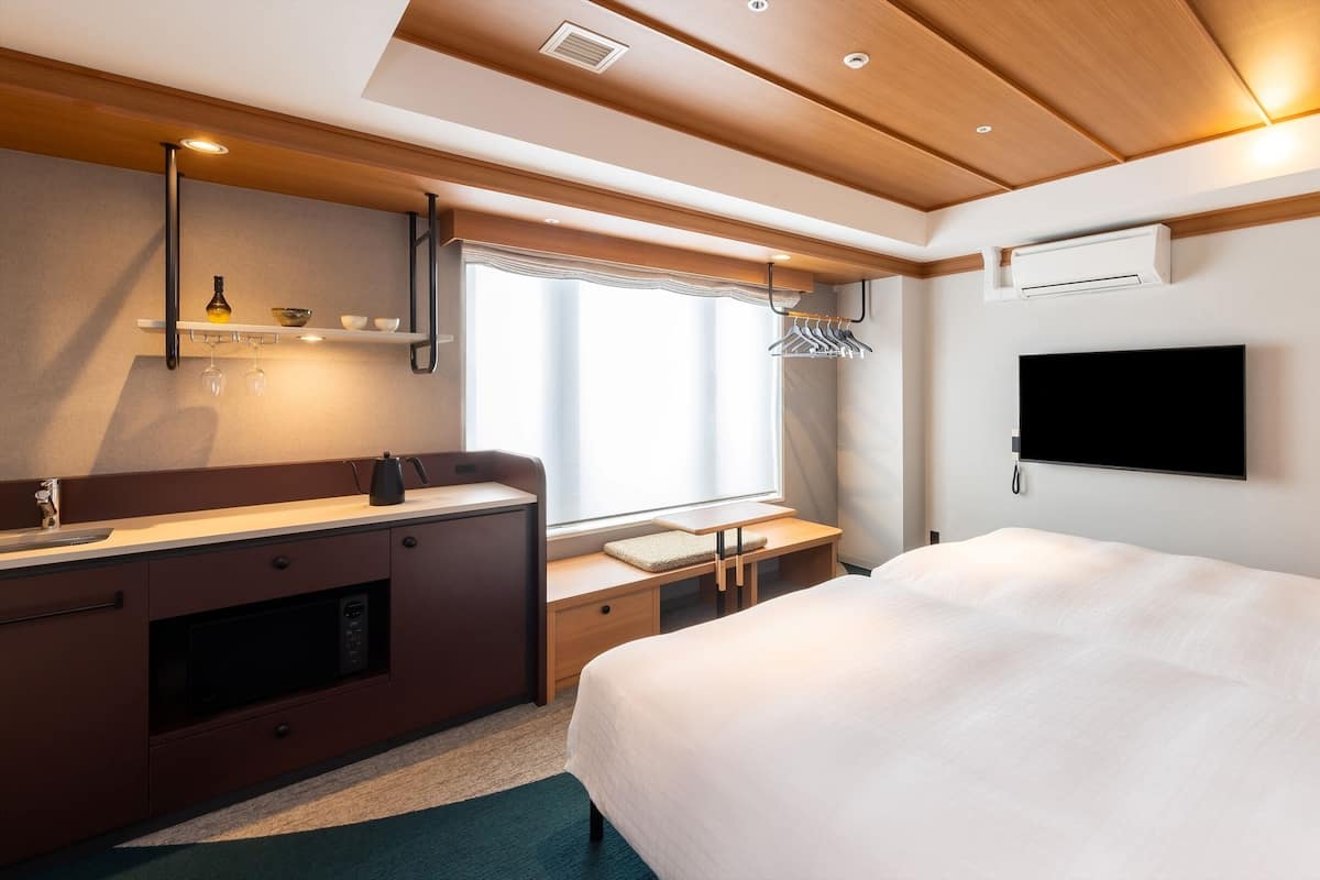 A modern hotel room with a wooden ceiling, a flat-screen TV mounted on the wall, a small desk with a kettle, and a large bed with white bedding.