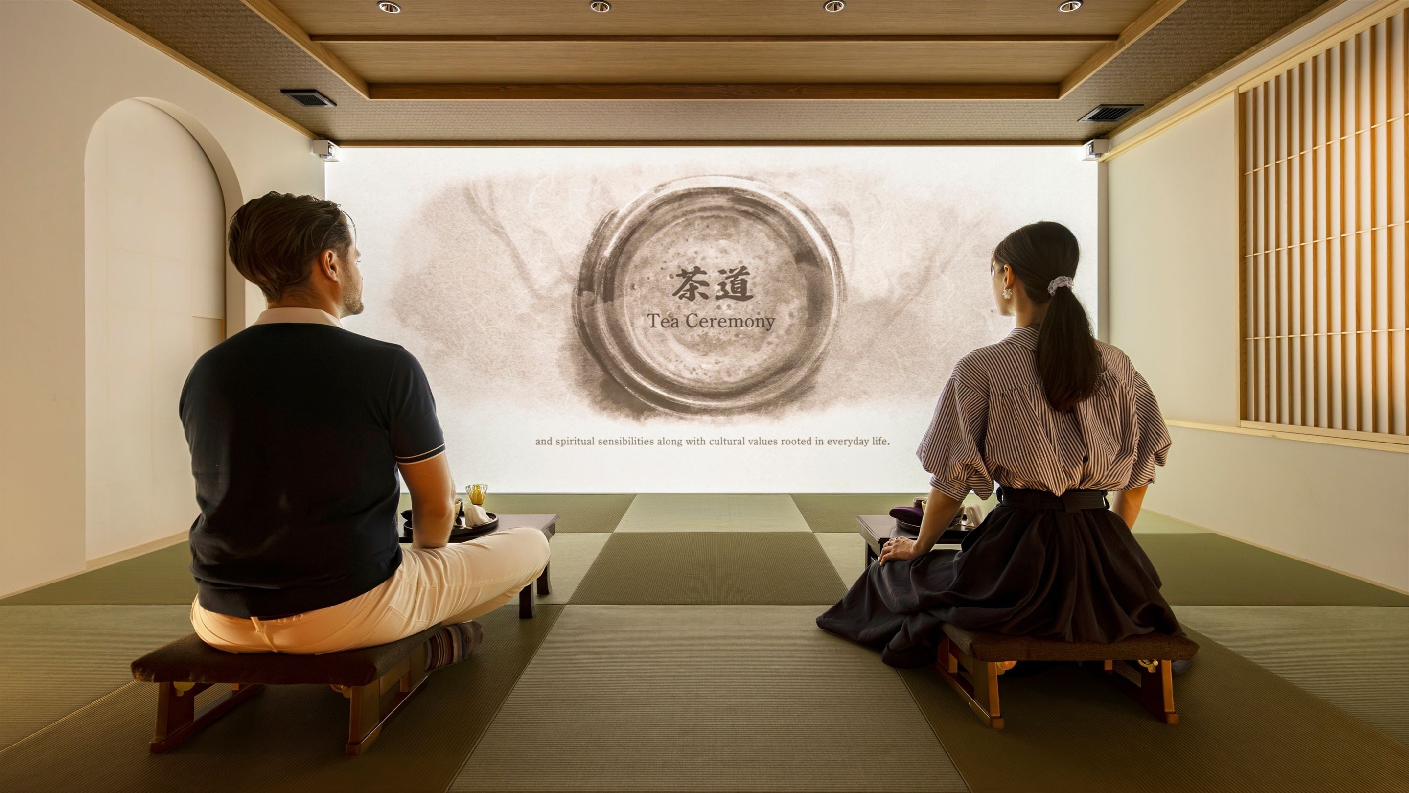 Dos personas sentadas sobre esteras de tatami frente a una pantalla que proyecta una ceremonia del té.