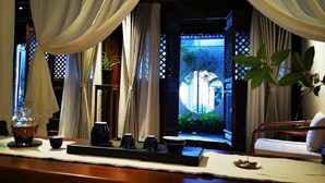 Une chambre avec un lit, une table avec un service à thé, et une vue sur un jardin par une fenêtre ornée de rideaux.