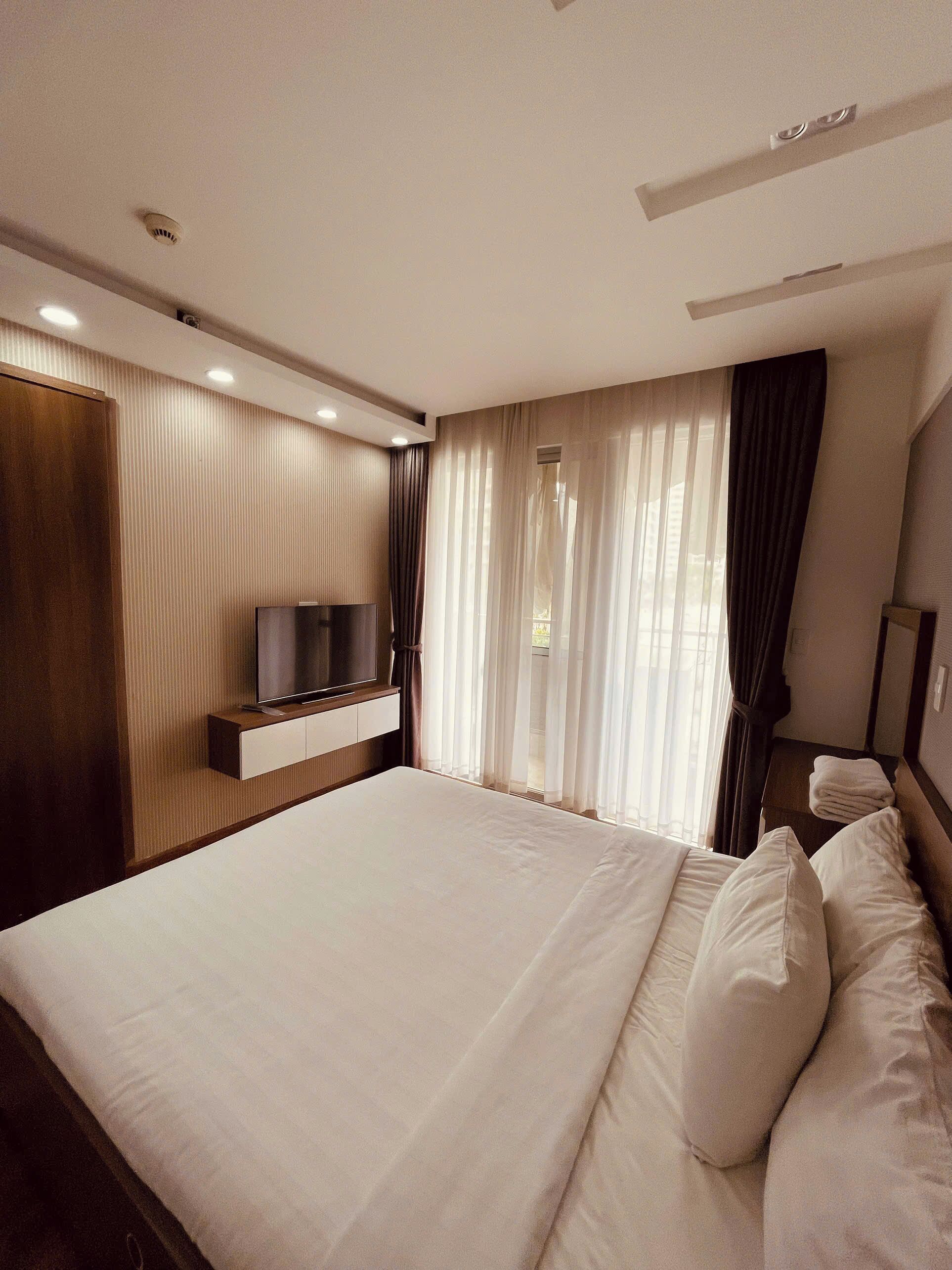 A modern hotel room with a large bed, a flat-screen TV mounted on the wall, and a window with sheer curtains.