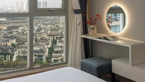 A hotel room with a large window offering a city view, a bedside table with a vase and book, and a bed with a red apple on the pillow.