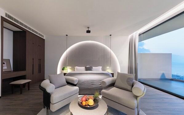 A modern bedroom with a curved ceiling, a bed, two sofas, a coffee table with fruit, and a view of the sea.