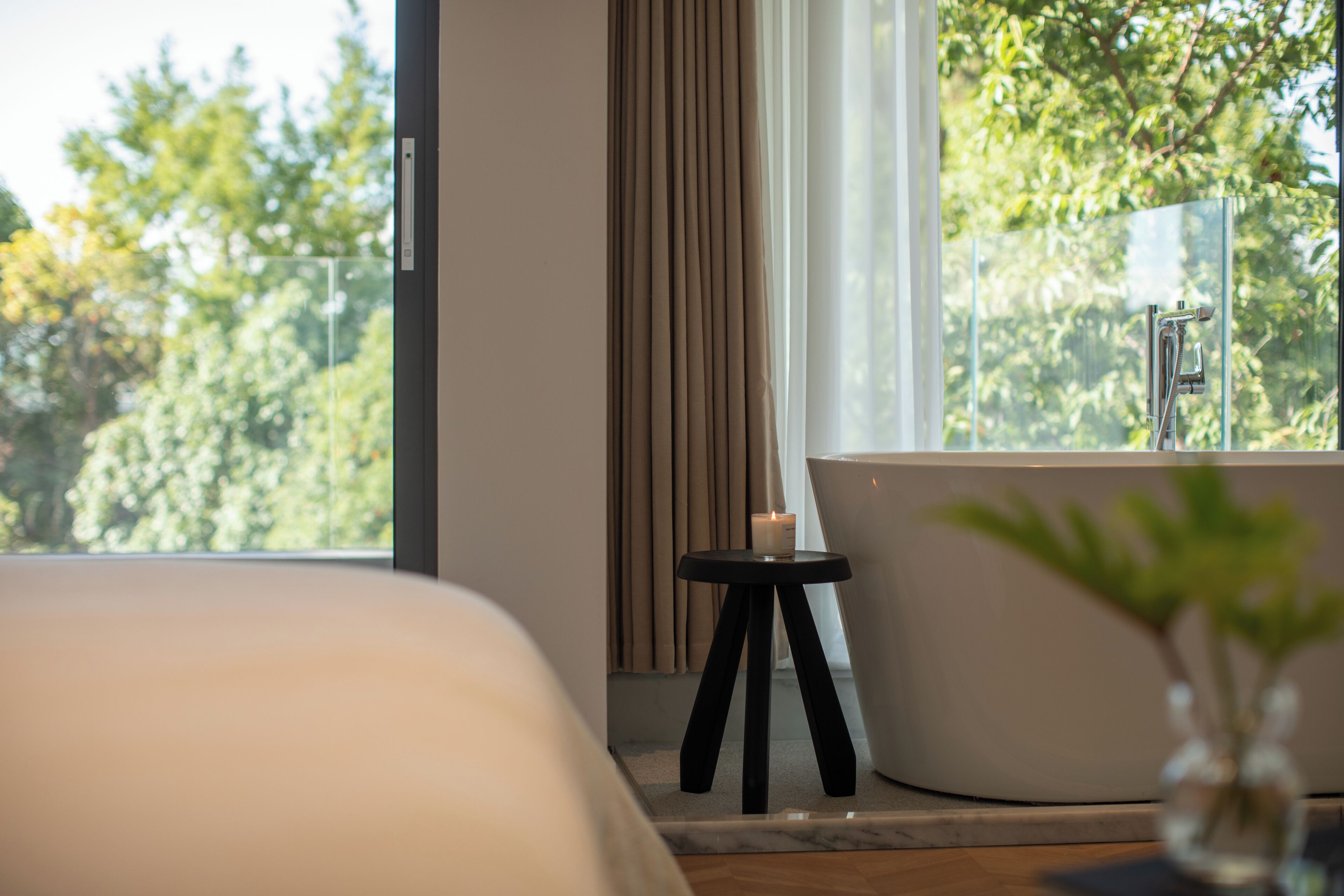 Une salle de bain moderne avec une baignoire, une petite table avec une bougie et une vue sur les arbres à travers une grande fenêtre.