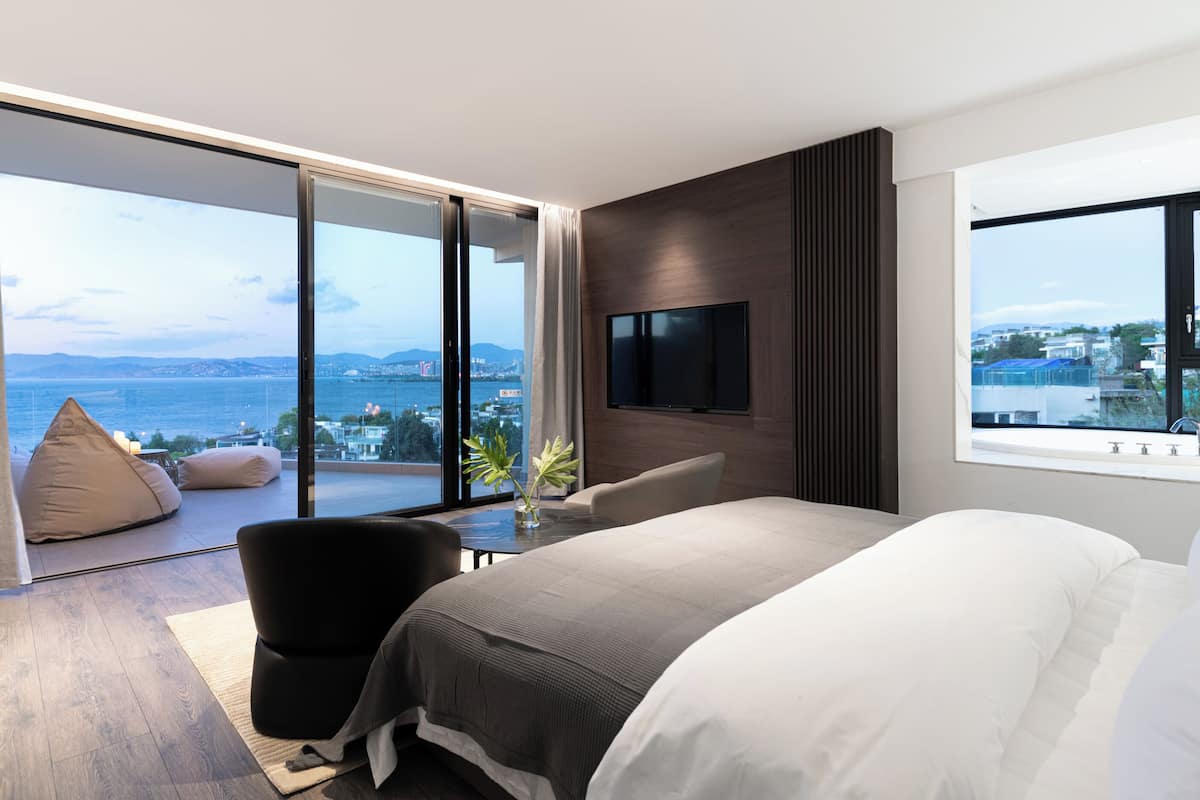 A modern bedroom with a large bed, a seating area with a chair, a TV mounted on the wall, and a view of the sea and cityscape.