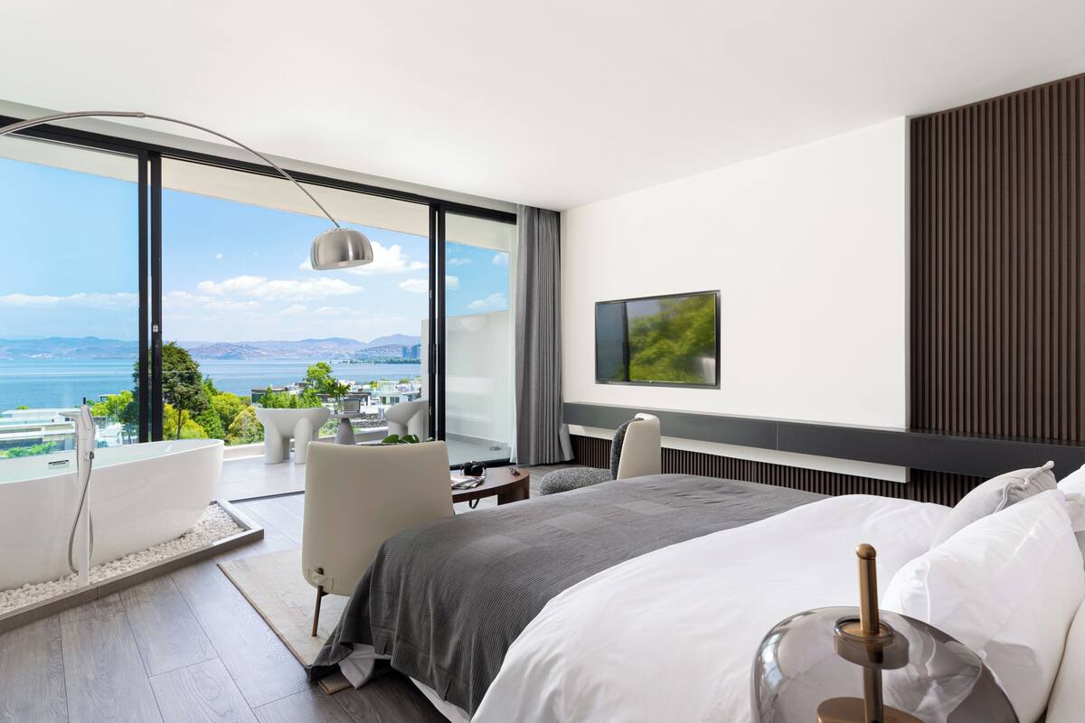 A modern hotel room with a large balcony, a bed, a desk, and a TV.