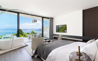 A modern hotel room with a large balcony, a bed, a desk, and a TV.