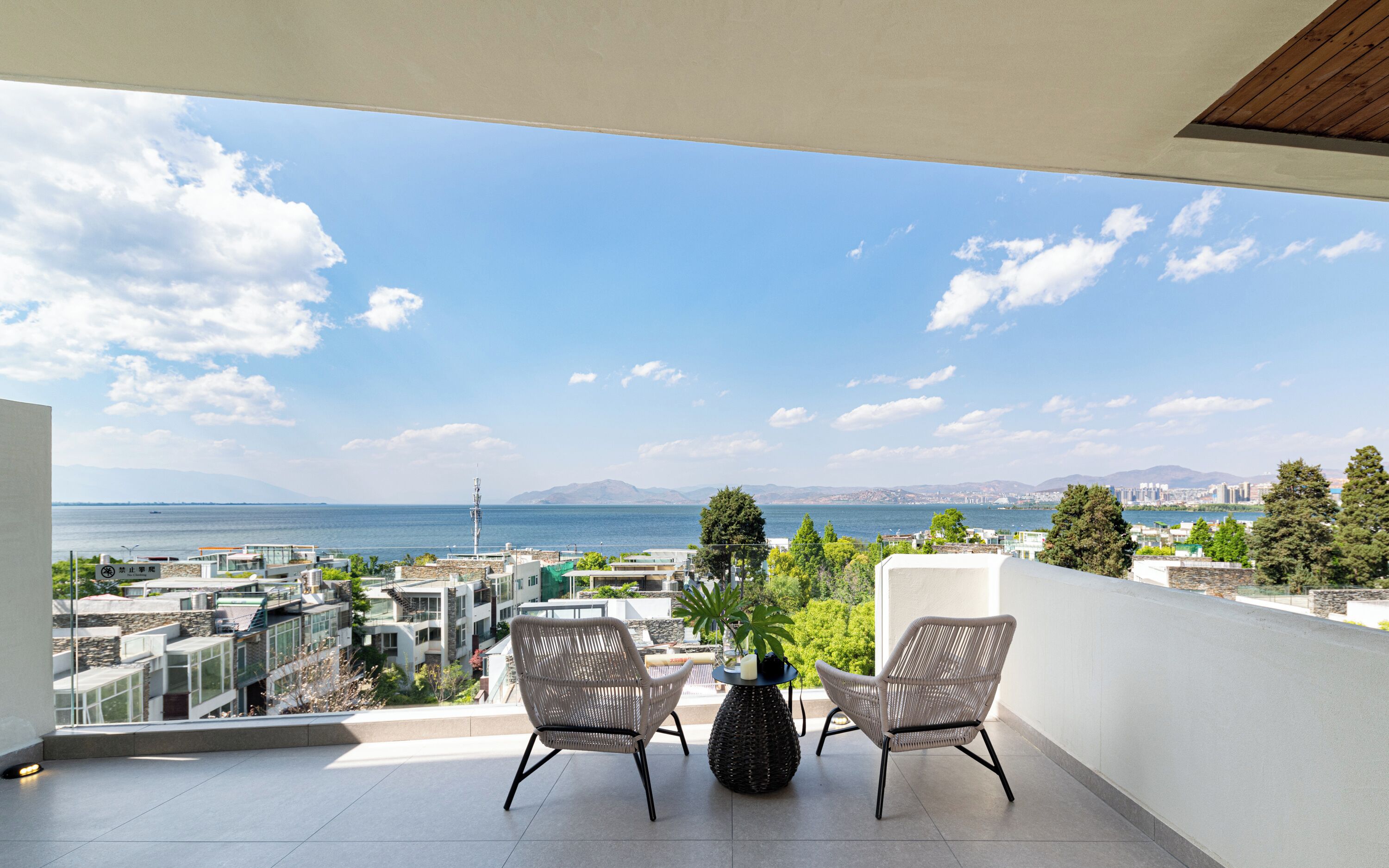 A balcony with two wicker chairs and a small table, overlooking a cityscape and a body of water.