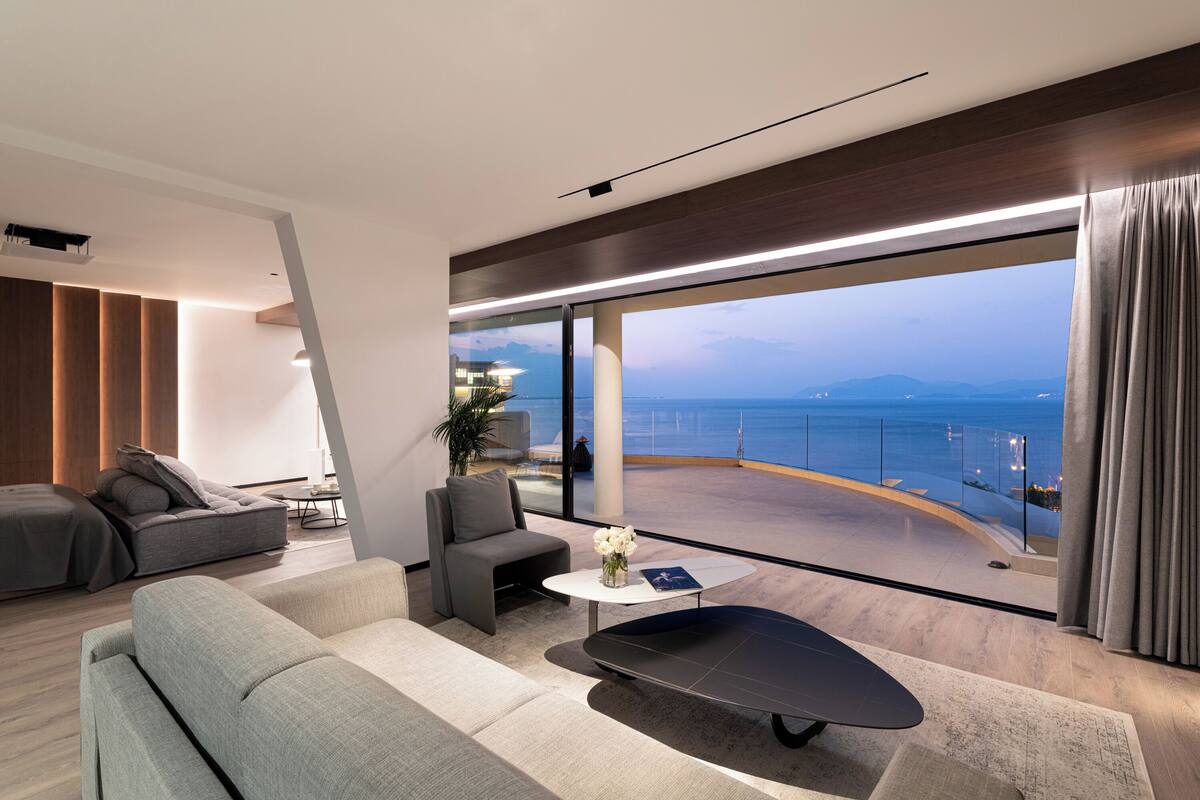 A modern living room with a sofa, armchair, and coffee table, a large window with a view of the sea, and a balcony with a railing.