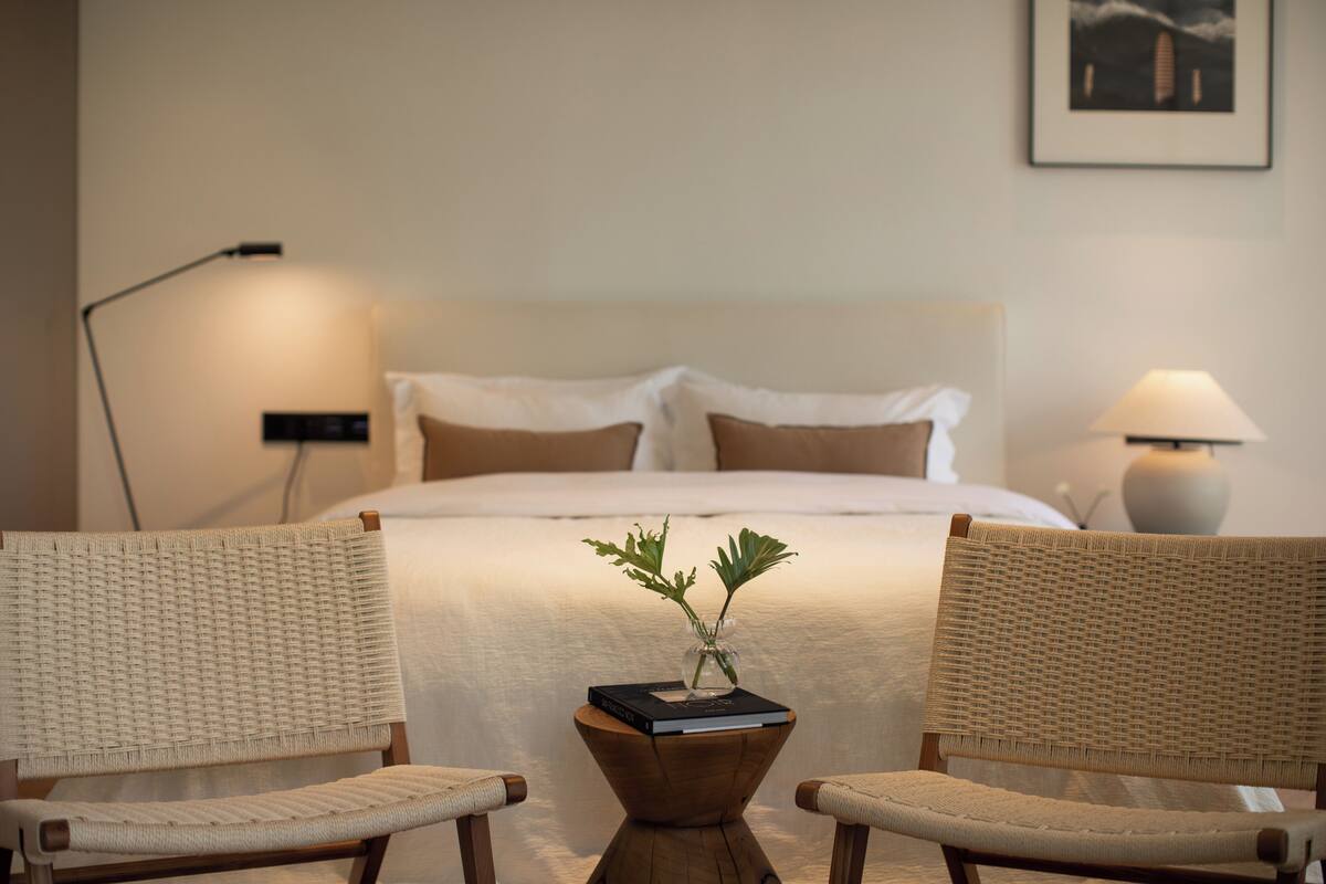 A hotel room with a bed, two wicker chairs, a small wooden table with a vase, and two bedside lamps.