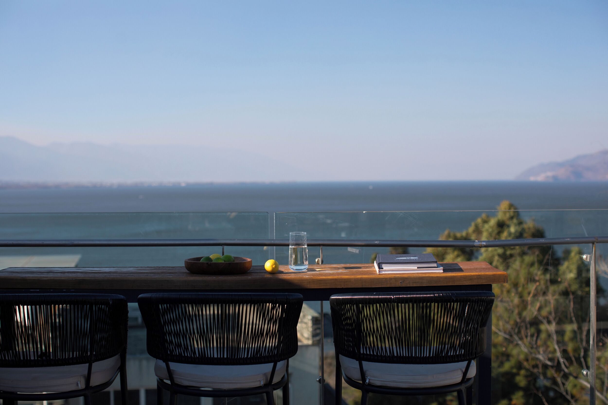 Ein Balkon mit einem Holztisch, zwei Korbstühlen, einer Obstschale, einem Glas Wasser und einem Buch.