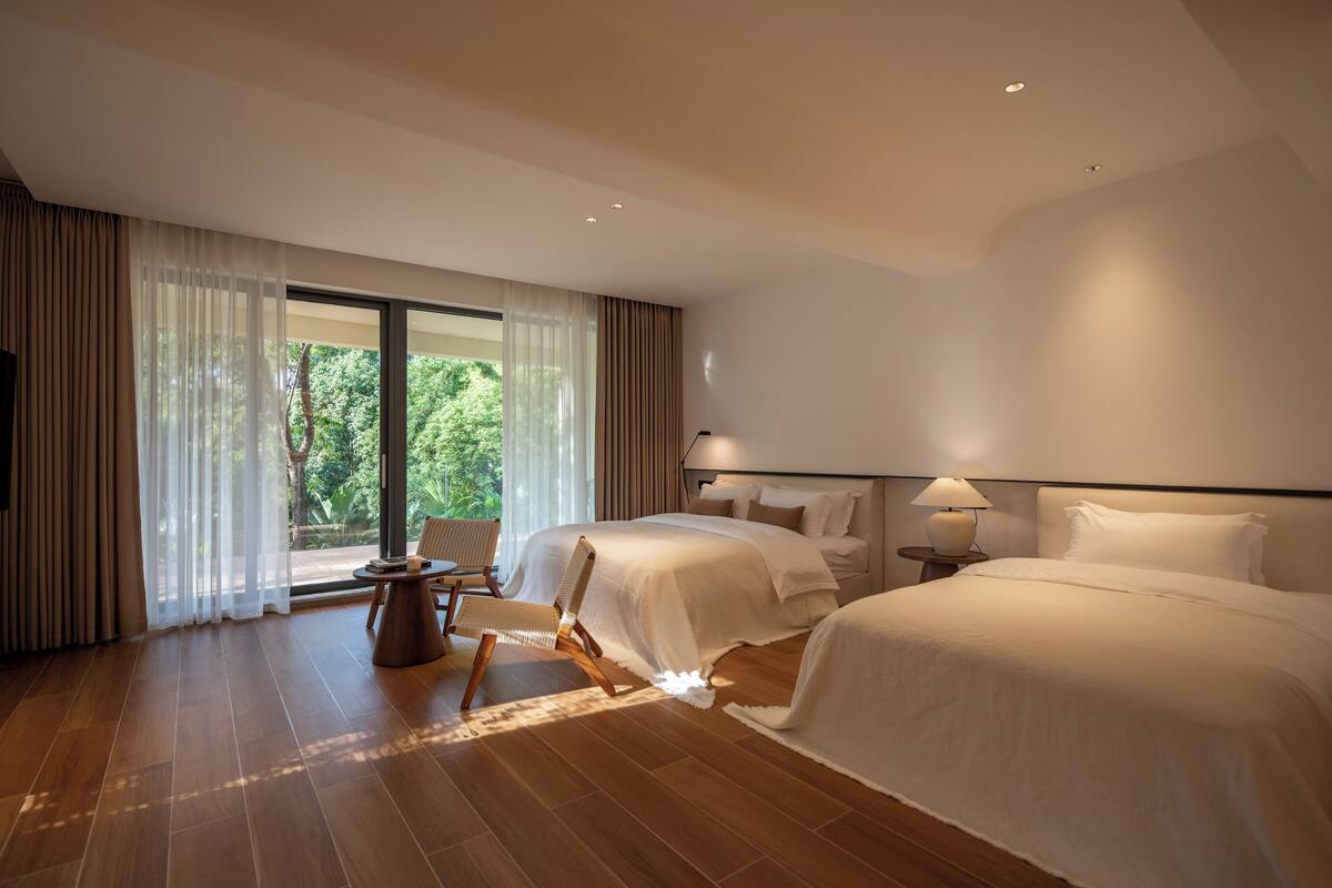 A hotel room with two beds, a wooden floor, a large window with curtains, a chair, and a nightstand with a lamp.