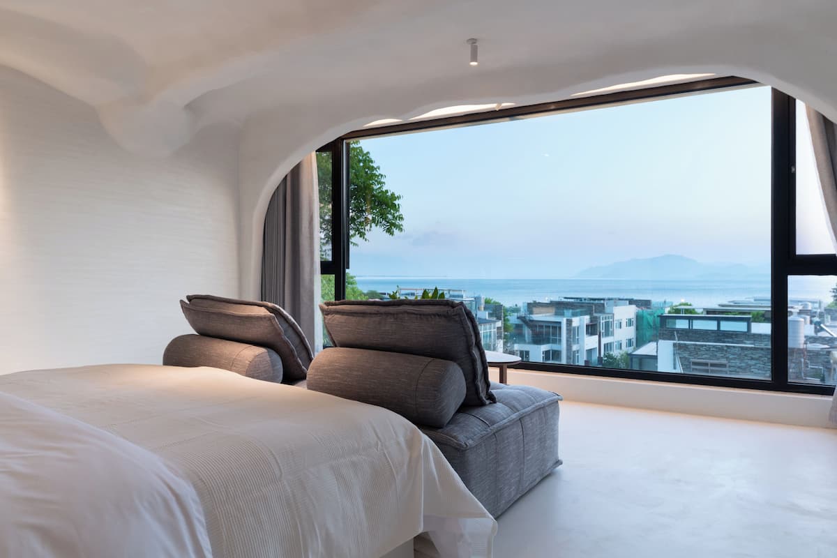 A modern bedroom with a large bed, two gray sofas, and a large window offering a city view.
