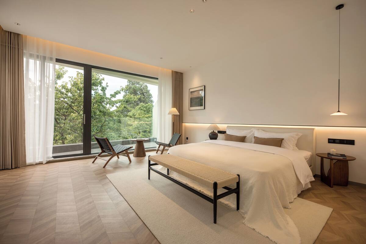 A modern bedroom with a large bed, a bench, a chair, a bedside table, and a view of greenery through large windows.