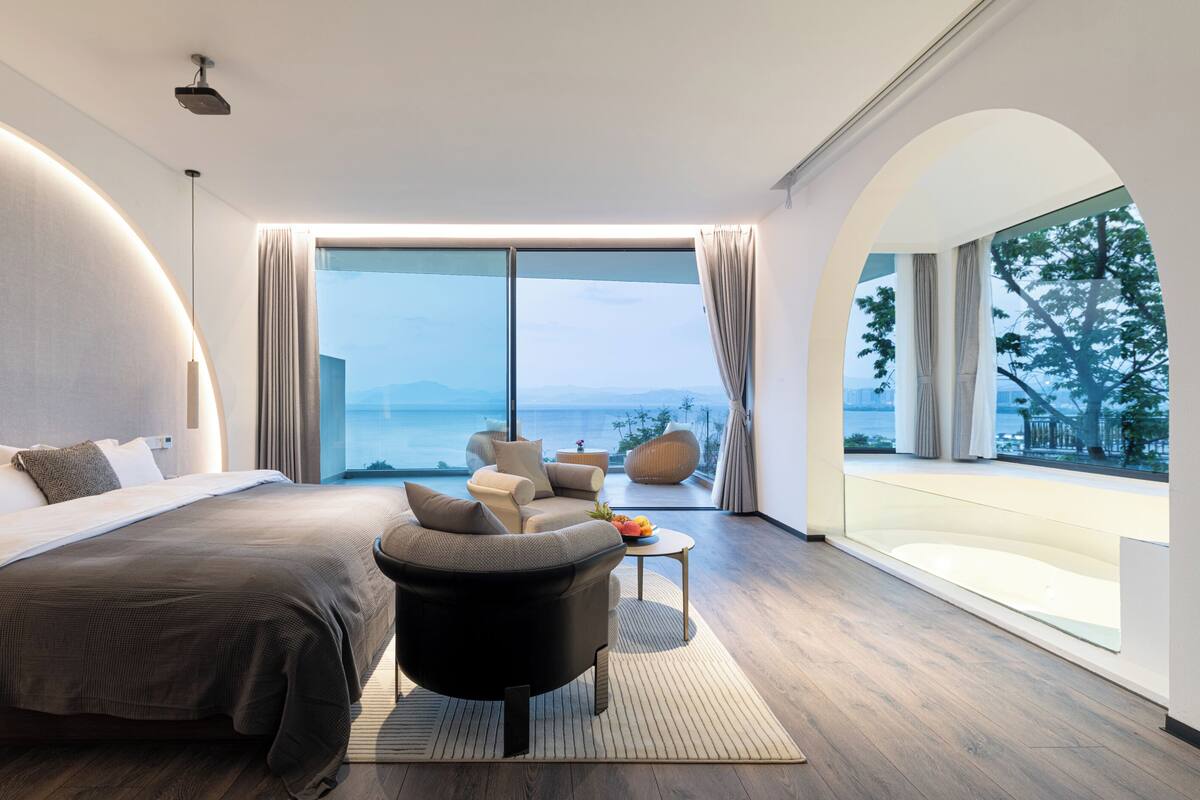 A modern bedroom with a large bed, a seating area with a small table, and a view of the sea.