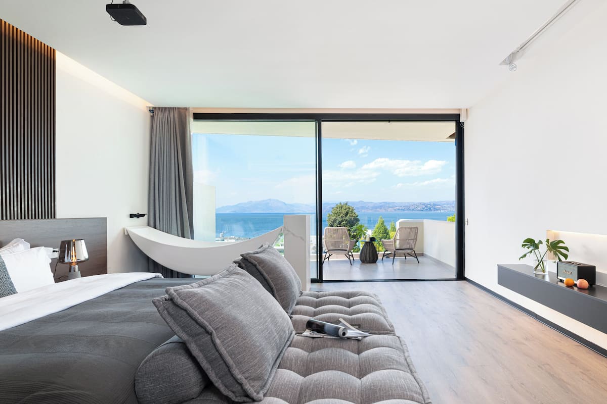 A modern bedroom with a large bed, a sofa, and a balcony with a view of the sea.