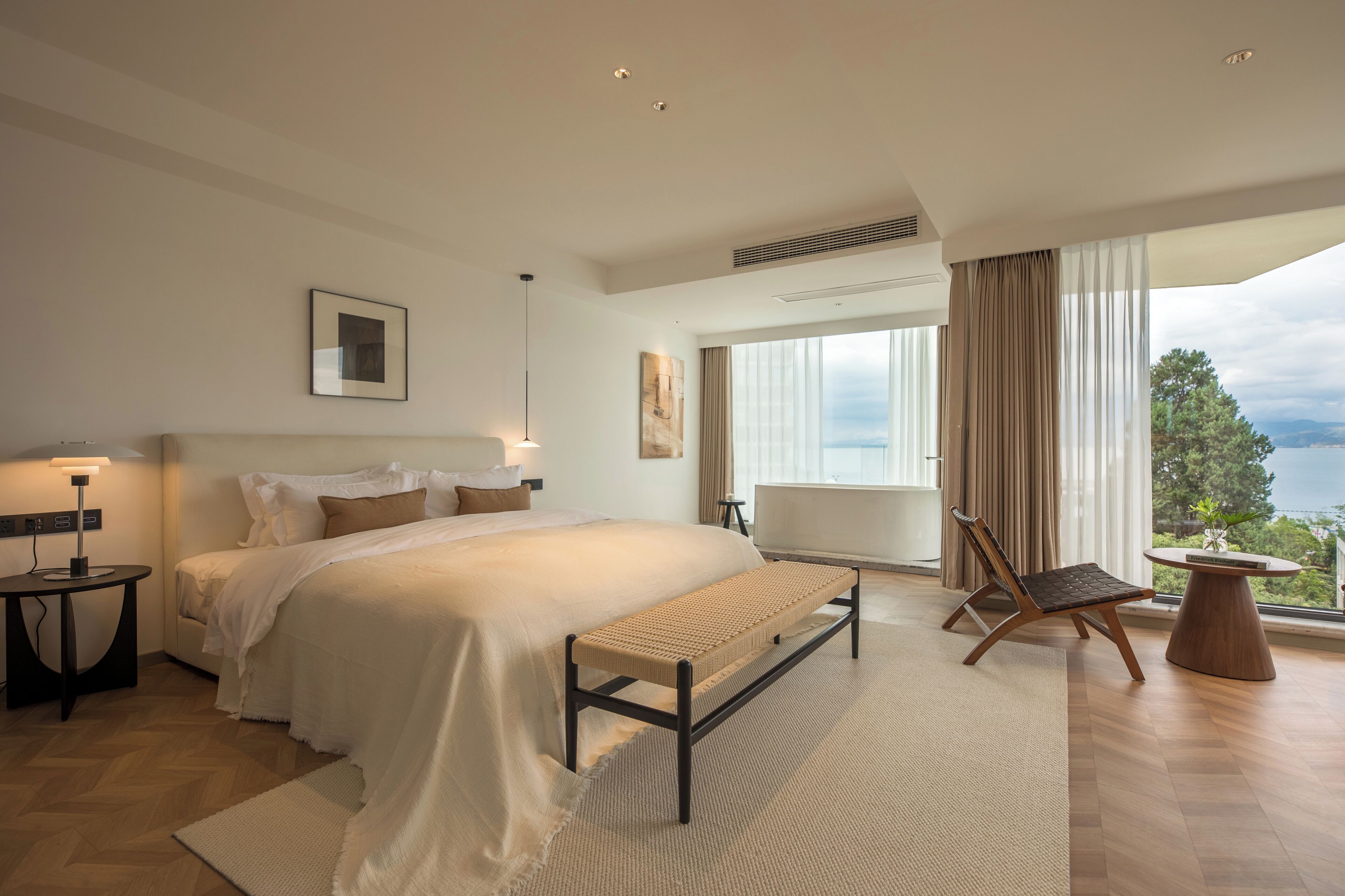 A modern bedroom with a large bed, a bench, a chair, a table, and a view of the outdoors.