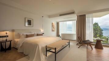 A modern bedroom with a large bed, a bench, a chair, a table, and a view of the outdoors.