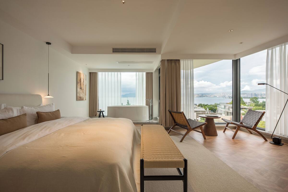A modern hotel room with a large bed, a sitting area with chairs, a small table, and a view of the city through large windows.