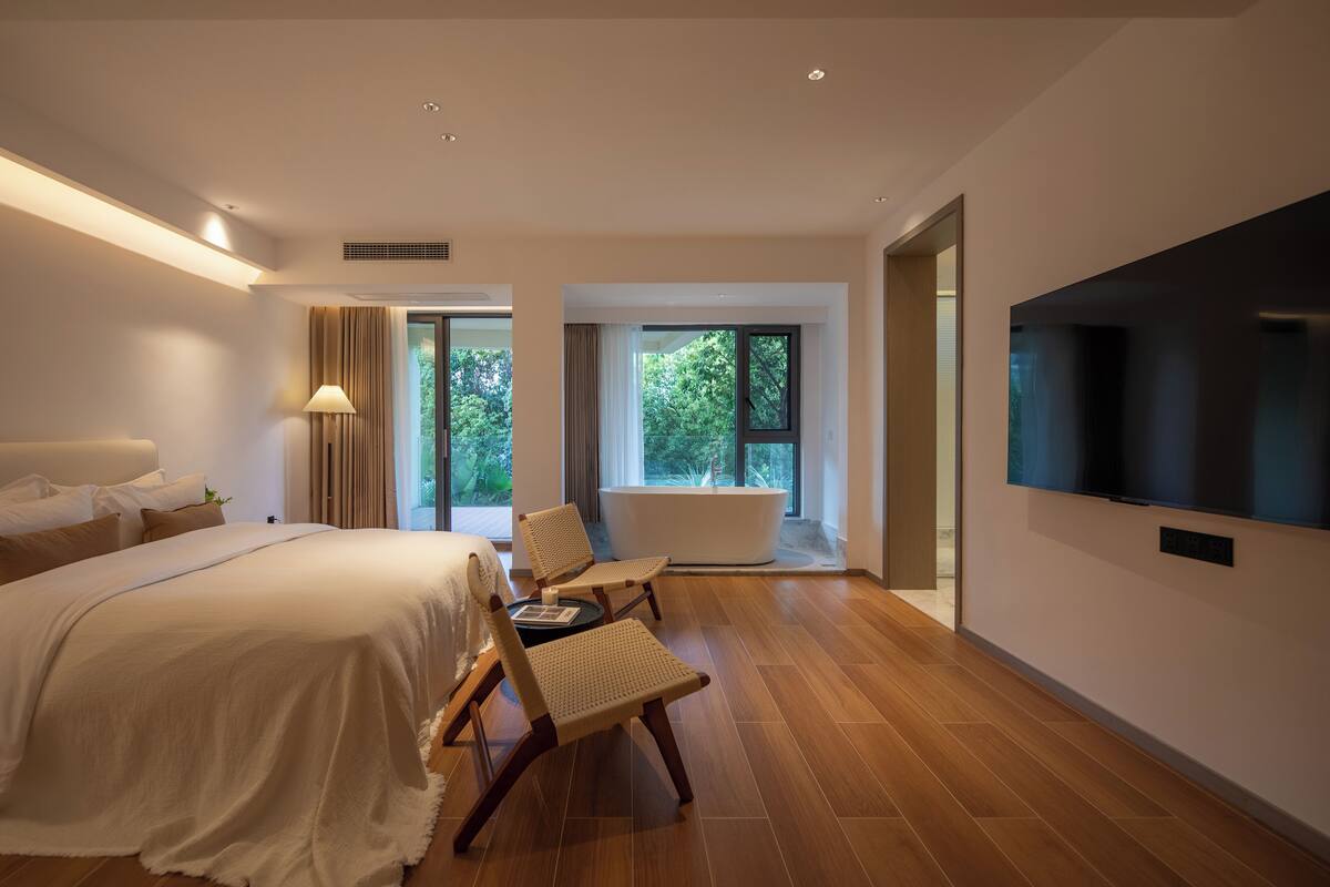 A modern hotel room with a large bed, a sitting area with chairs, a bathtub, and a TV mounted on the wall.