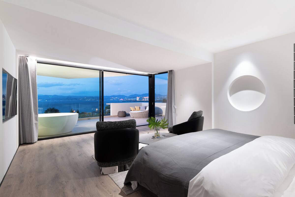 A modern hotel room with a large bed, a seating area with a view of the sea, and a circular light fixture on the wall.
