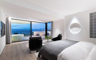 A modern hotel room with a large bed, a seating area with a view of the sea, and a circular light fixture on the wall.