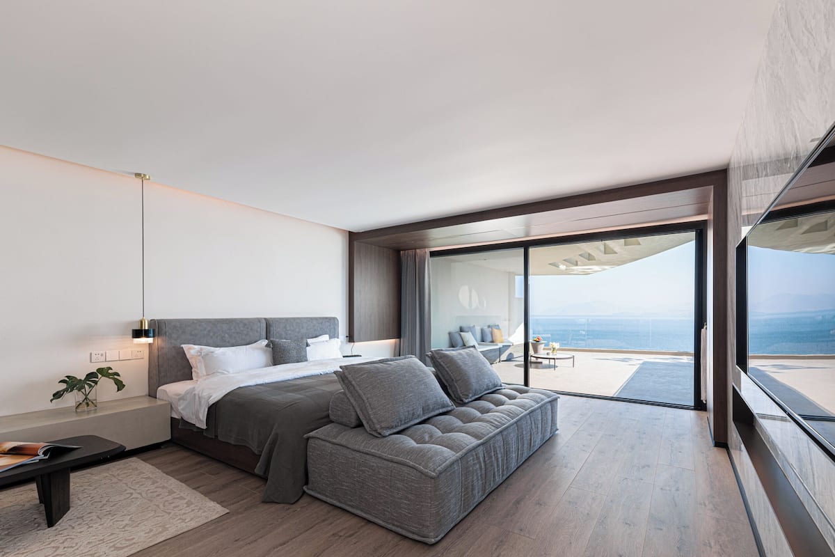 A modern bedroom with a large bed, a grey sofa, and a balcony with a view of the sea.