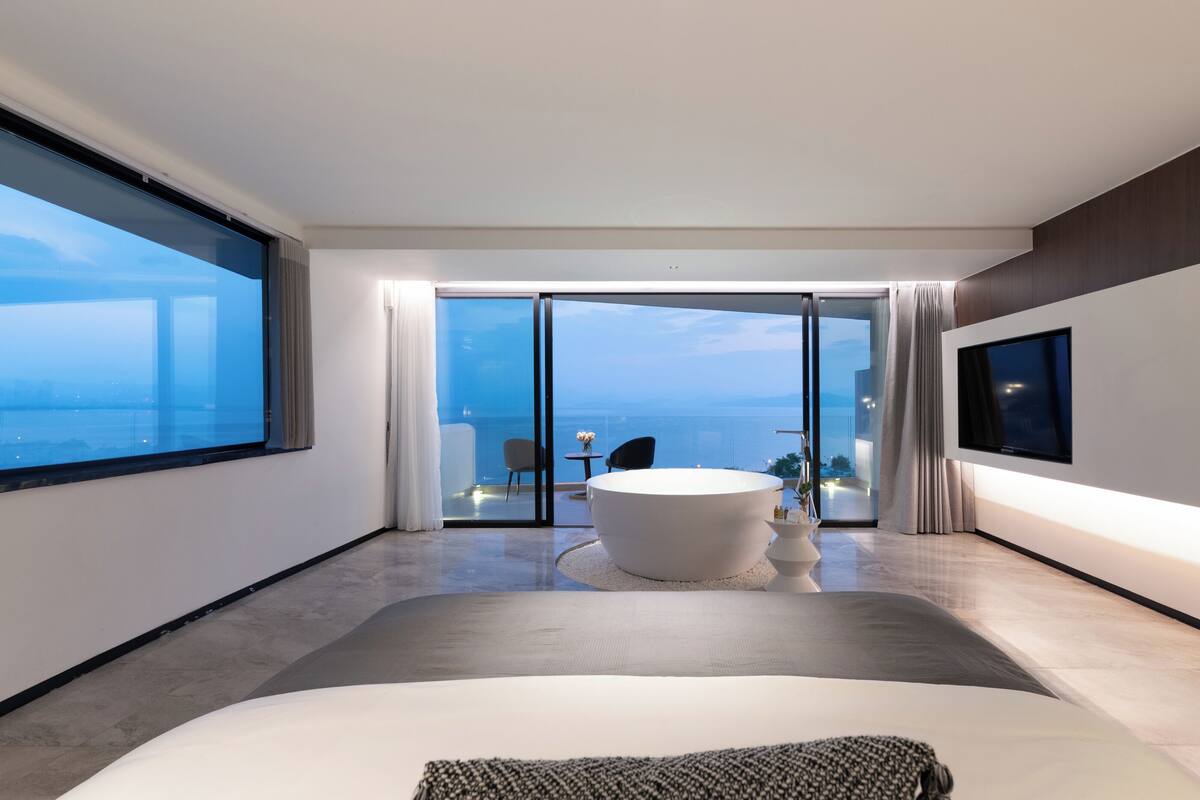 A modern hotel room with a large bed, a bathtub, a TV, and a balcony with a view of the sea.