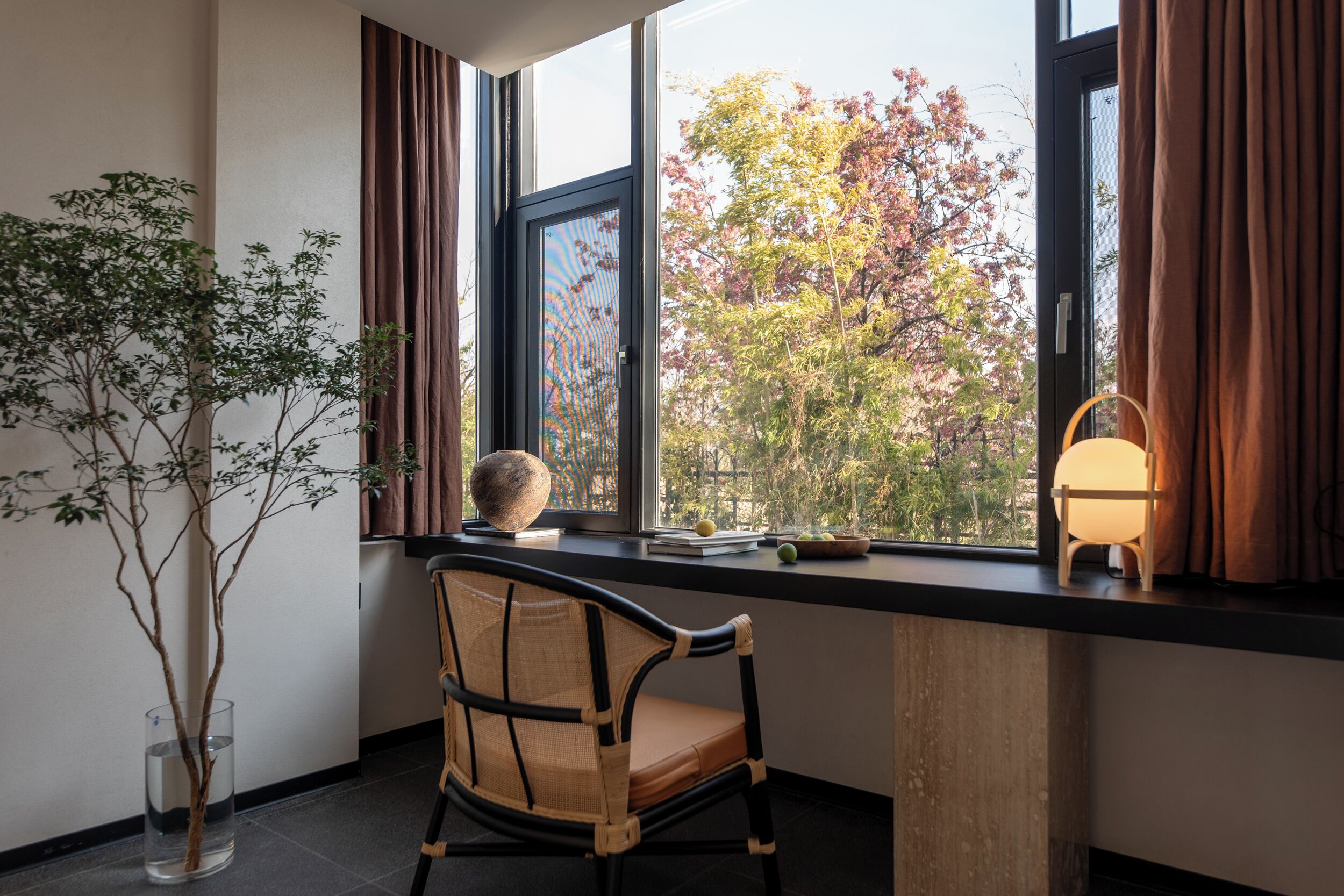 A room with a large window, a chair, a small table with a lamp, and a potted plant.