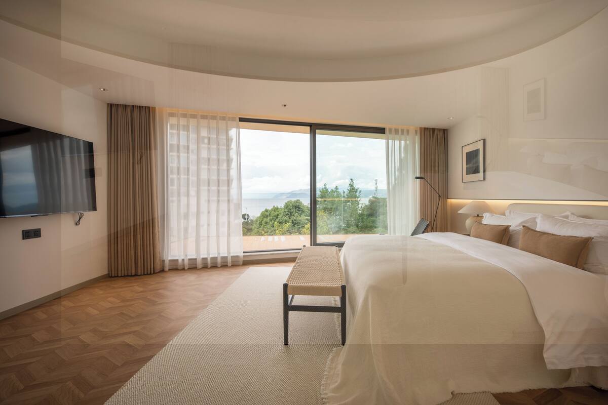 A modern hotel room with a large bed, a flat-screen TV, a balcony with a view, and a large window with curtains.