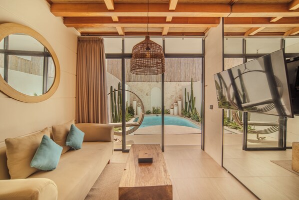 A modern living room with a sofa, coffee table, and a large mirror. There is a pool visible through the glass doors.