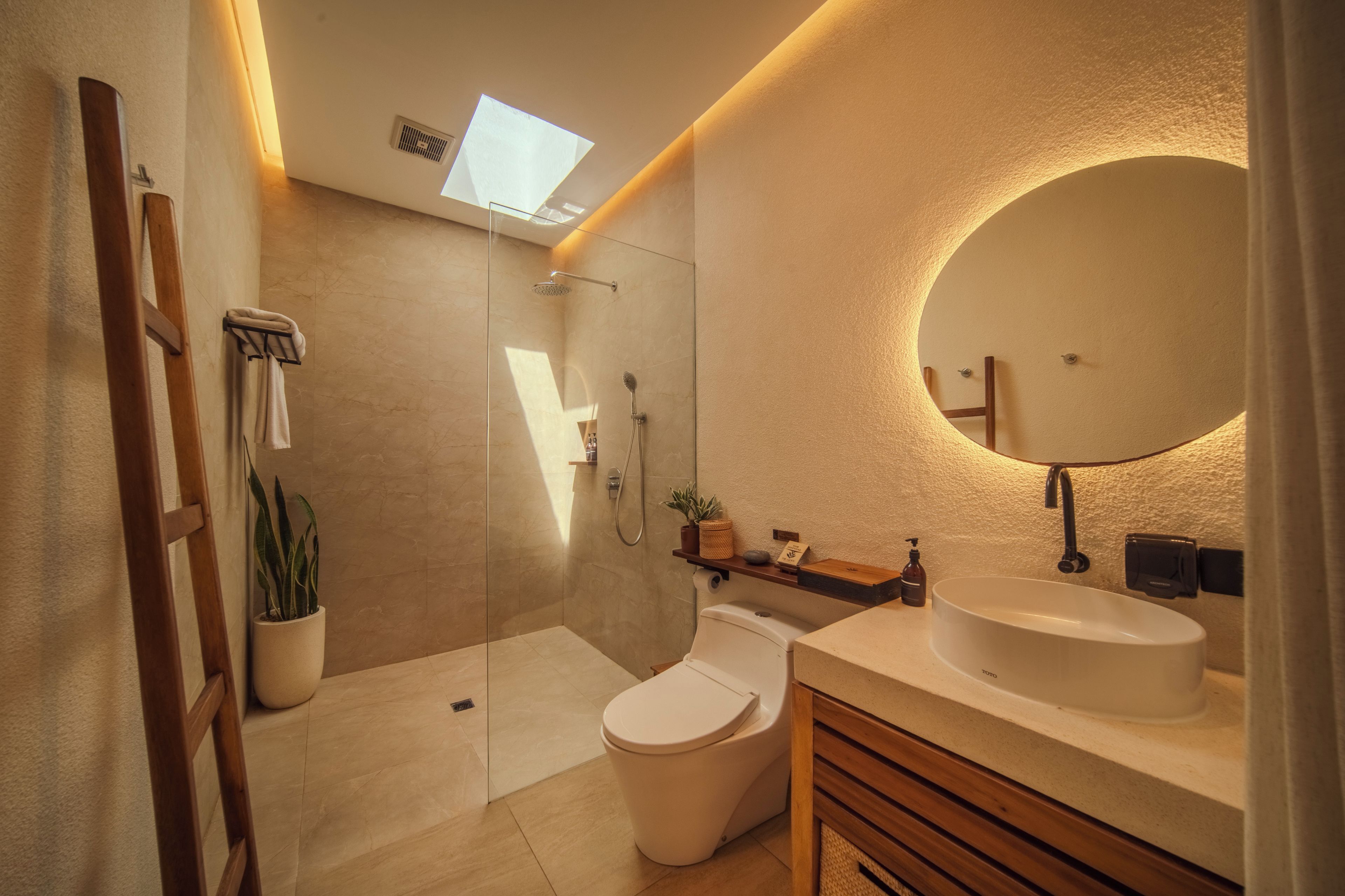 A modern bathroom with a glass shower enclosure, a toilet, a sink with a round mirror, and a skylight.