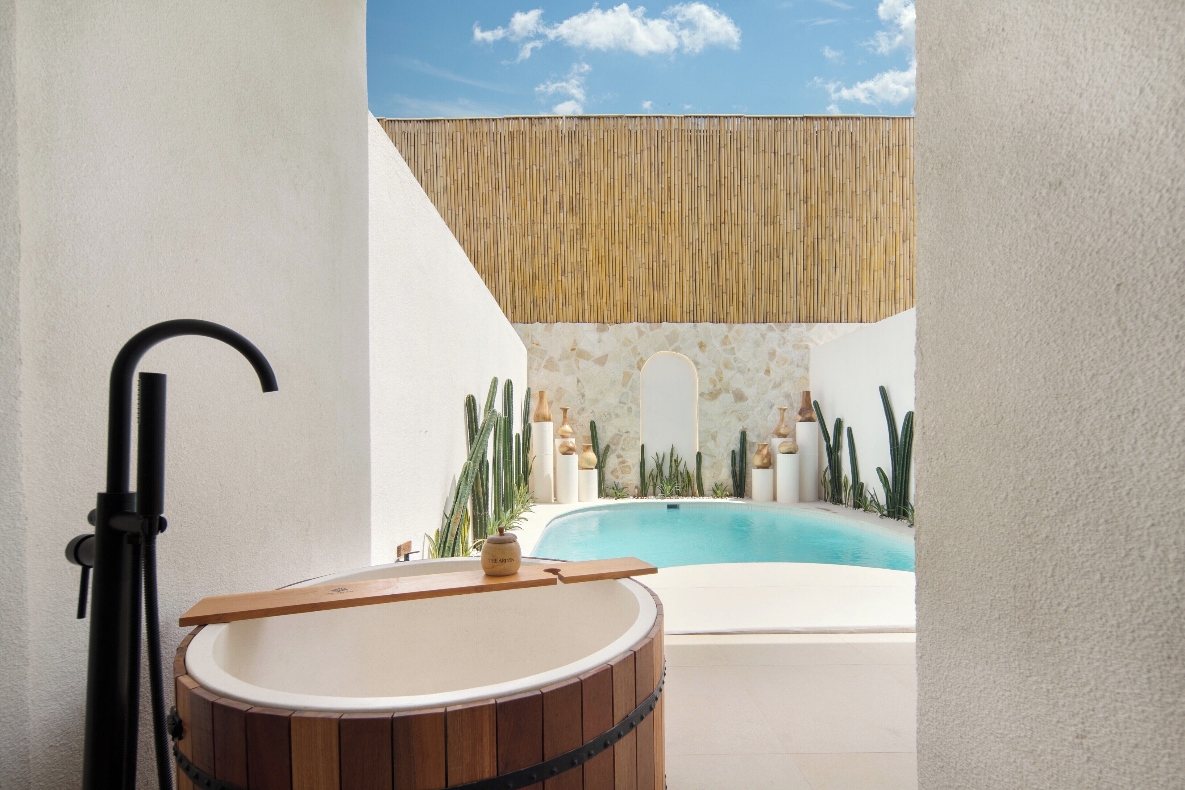 A spa area with a wooden bathtub, a hot tub, and a pool, surrounded by plants and a bamboo wall.