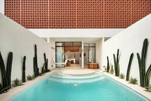 Un área de piscina interior moderna con un jacuzzi, rodeada de cactus y plantas, y vistas hacia una habitación bien iluminada con una cama y un armario de madera.