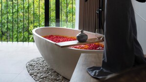 Una bañera con pétalos de flores rojos y amarillos, un lavabo de piedra y un vaso en un balcón con vistas a la vegetación.