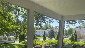 En overdækket terrasse med et bord og stole, et mønstret tæppe og udsigt til et boligområde med huse og en parkeret campingvogn.