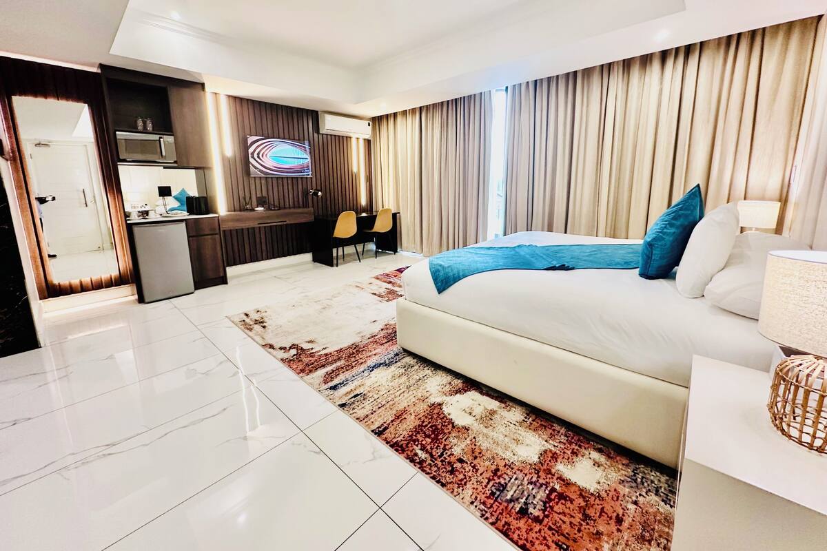 A modern hotel room with a large bed, a desk with a chair, a small kitchenette, and a bathroom.