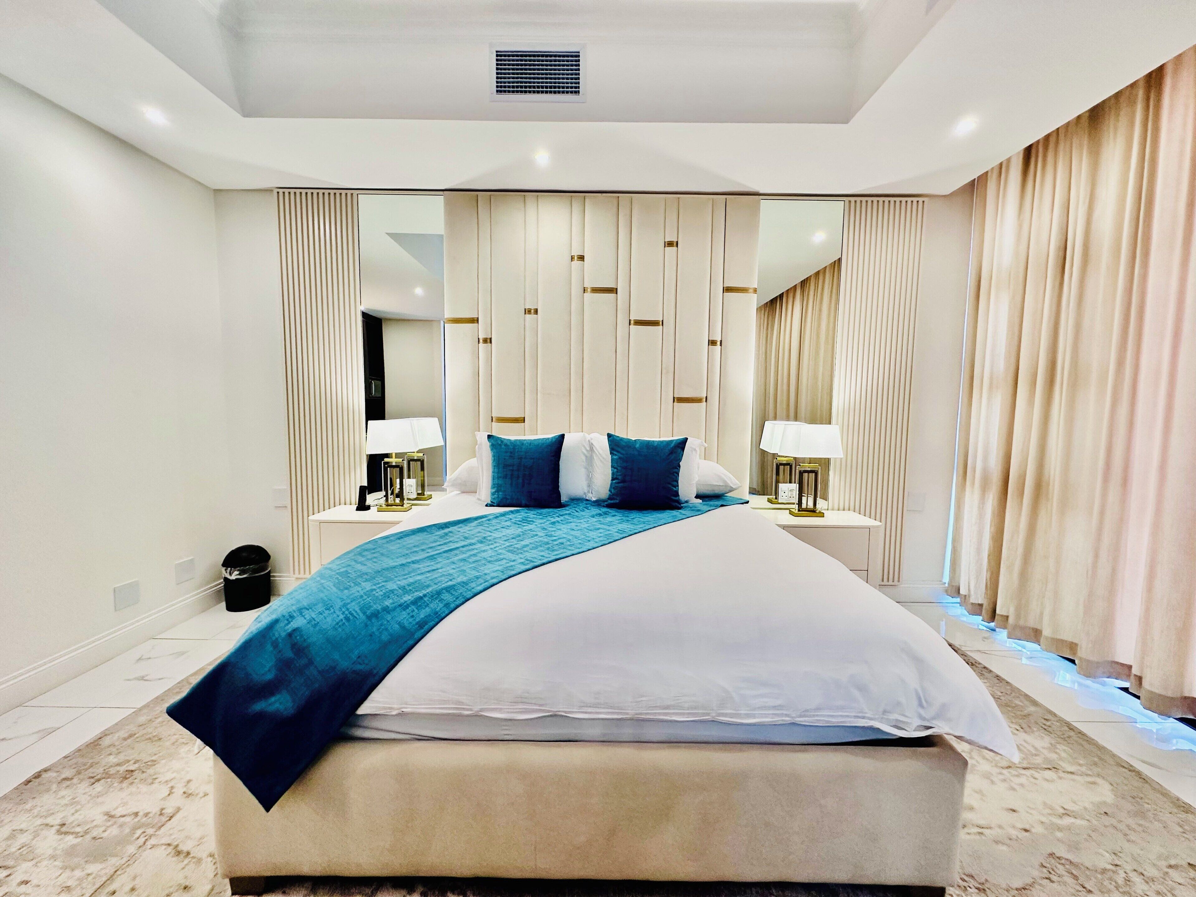 Une chambre d’hôtel moderne avec un grand lit, deux tables de chevet avec des lampes, un grand miroir et un rideau à la fenêtre.