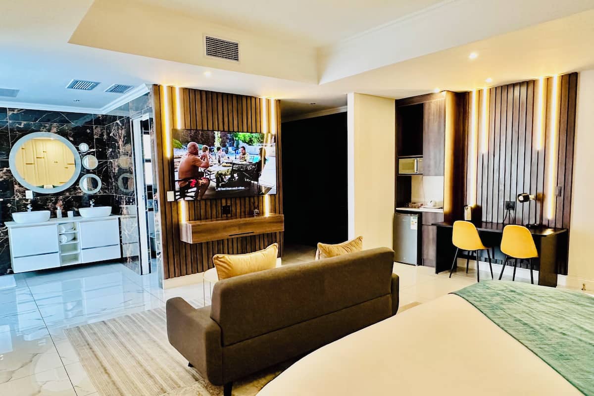 A modern hotel room with a large bed, a sofa, a TV, a kitchen area, and a bathroom with a vanity and mirror.