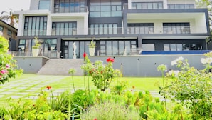 Um edifício moderno de vários andares, com varandas, entrada central e um jardim bem cuidado, com diversas plantas e flores.