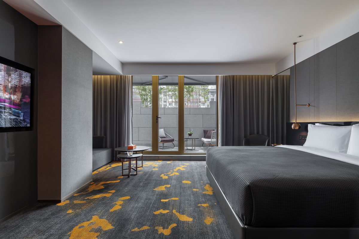 Una camera d'hotel con un letto grande, una TV a parete, un tavolino con una sedia e vista all'esterno attraverso ampie finestre.