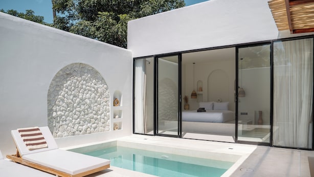 A modern outdoor pool area with lounge chairs, a stone wall, and a glass-enclosed bedroom with a bed and nightstands. - Villa Kasih Satu (Gili Trawangan)