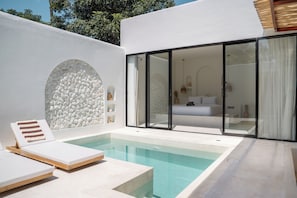 Uma área de piscina moderna ao ar livre com espreguiçadeiras, um muro de pedra e um quarto com cama e mesas de cabeceira, protegido por vidro.