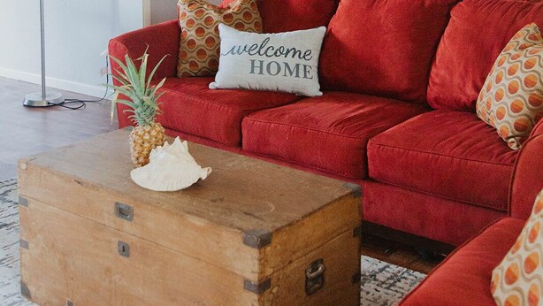 Ein rotes Sofa mit einem 'welcome home'-Kissen, eine Holzablage und eine Stehlampe in einem Wohnzimmer mit einem Wandbild von einem Sonnenuntergang.