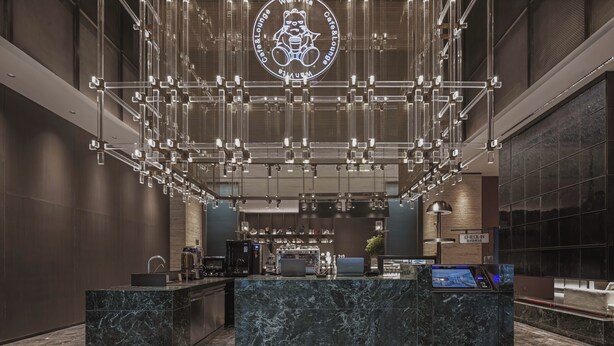 Un hall d’hôtel moderne avec un comptoir d’accueil en marbre, un kiosque d’enregistrement numérique et un grand logo lumineux sur le plafond.