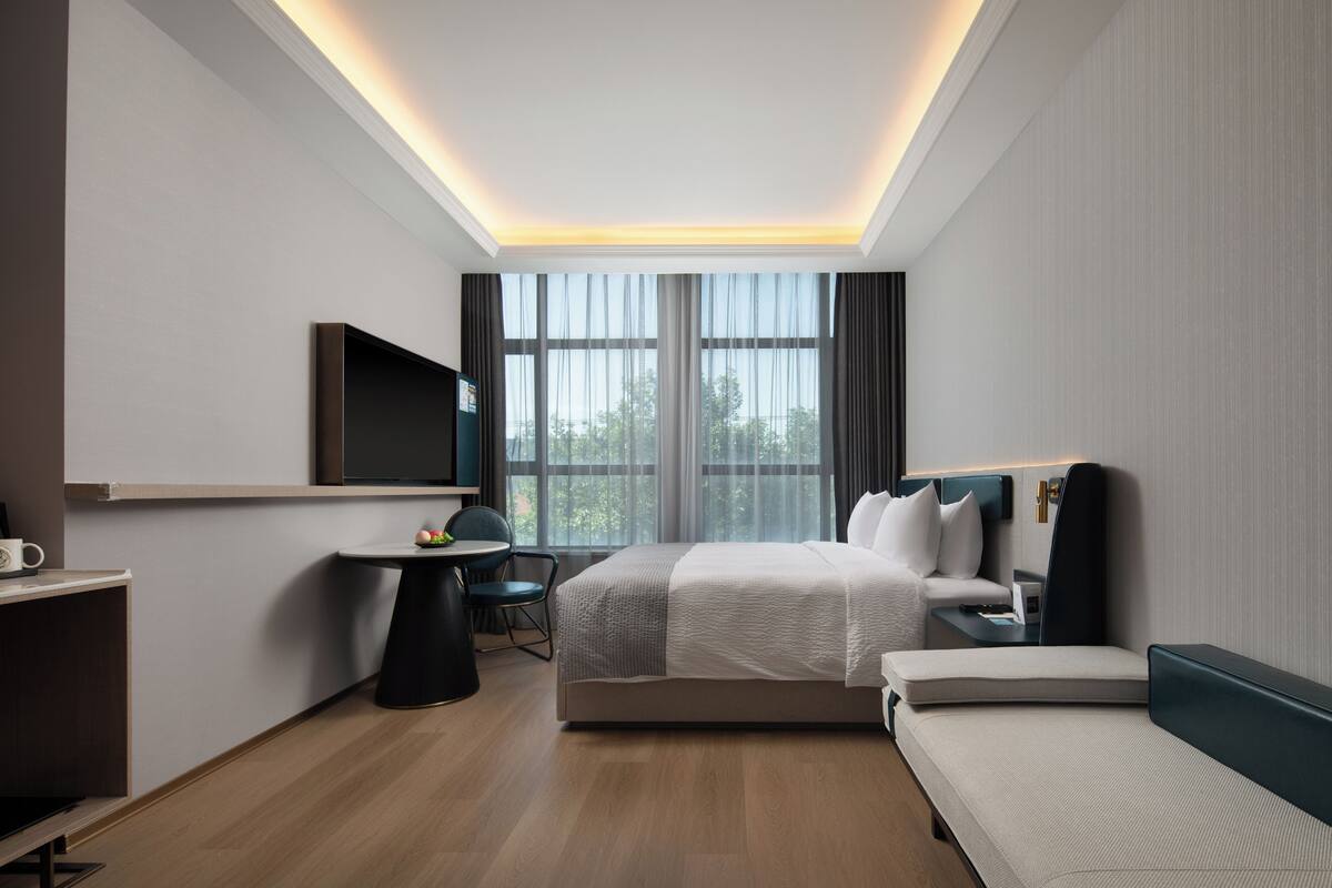 A modern hotel room with a bed, bedside table, TV, and a desk with a chair.
