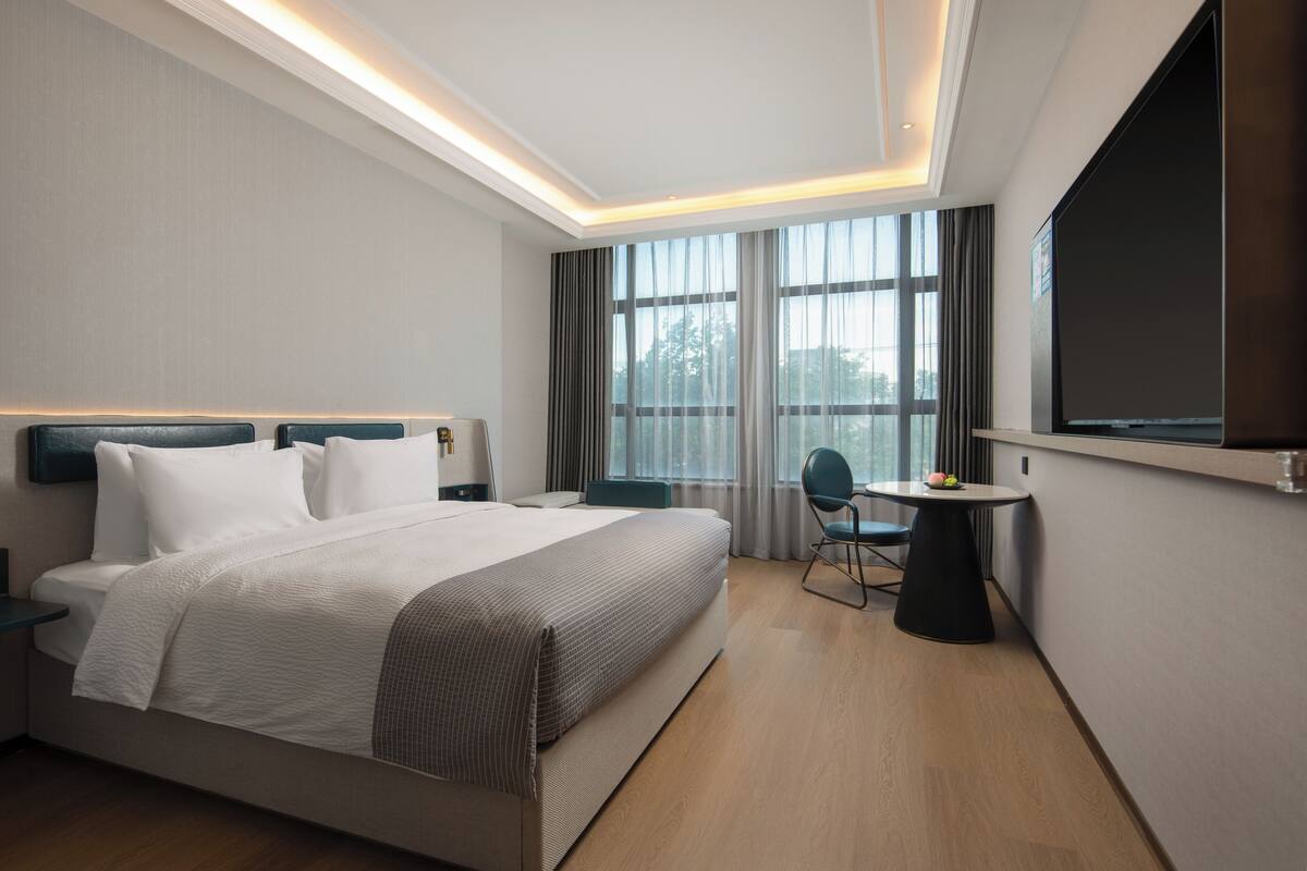 A modern hotel room with a large bed, a small round table, a chair, and a TV mounted on the wall.