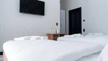 A hotel room with two beds, a flat-screen TV mounted on the wall, a wooden side table with toiletries, and a dark door.