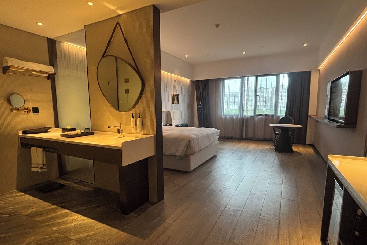 A modern hotel room with a large bed, a separate sitting area, a bathroom with a large mirror, and a television.